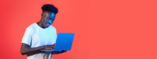 A young black man is engaged with his laptop, beaming with joy as he interacts with the screen. The solid red background creates a lively atmosphere, drawing attention to his focused expression. photo