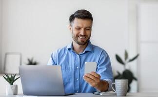 Handsome male office manager using smartphone at workplace, sitting at desk, checking social media or browsing app, answering to message, having break, looking at screen and smiling, copy space photo