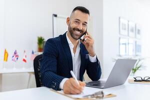 A well-dressed man sits at a desk, smiling while on a phone call. He writes notes on a clipboard, with a laptop open in front of him and flags from various countries displayed nearby. photo
