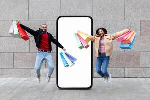 A young couple jumps in excitement while holding colorful shopping bags in front of a large blank smartphone. They convey joy and enthusiasm in their retail experience. photo