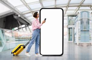 A traveler stands in an airport terminal, checking their phone while leaning against a large blank advertisement display. A yellow suitcase rests beside them, adding color to the scene. photo