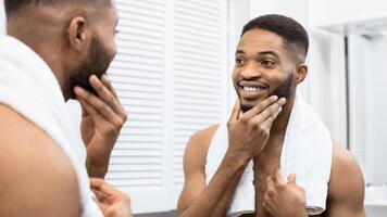 Happy afro man with bare torso touching beard looking in mirror at bathroom. Modern man in bathroom concept photo