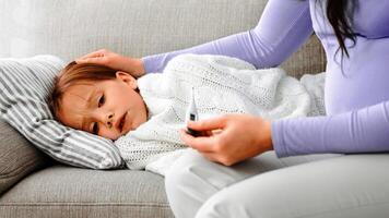 Expecting mother checking her ill daughter's temperature with a thermometer, girl laying on sofa, care concept, cropped photo