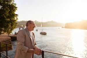 A smiling man leans on a railing by the river as the sun sets, casting a warm glow over the water and surrounding landscape. Boats gently sail by in the distance. photo