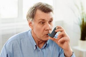 Senior Man Using Asthma Inhaler For Allergies In Modern Clinic, Copy Space photo