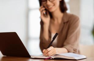Taking Notes. Young secretary sitting at desk with laptop computer and talking on mobile phone, writing in notebook photo