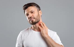 Middle aged bearded man scratching highlighted with red neck on grey studio background, copy space. Annoyed man suffering from itch, having rash on his neck. Allergic reaction, eczema concept photo