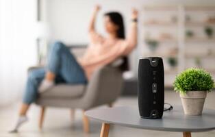 Modern Technology Concept. Closeup of smart portable wireless speaker on the table, selective focus. Young woman listening to music and dancing, sitting on armchair in the blurred background photo