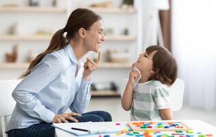 Speech training for kids. Professional woman specialist training with little boy at cabinet, teaching him right articulation exercises, side view photo