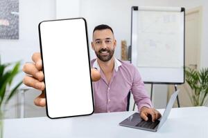 A smiling man sits at a desk in a contemporary office, holding a blank smartphone screen toward the viewer while working on a laptop, highlighting a friendly and tech-savvy atmosphere. photo