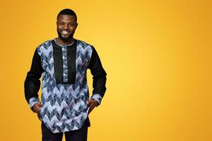 Cheerful black man in traditional african costume smiling over pink studio background, putting hands in pockets, copy space. Confident african american guy posing in ethnic colorful clothes photo