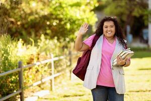 A cheerful student, dressed in a pink shirt and a vest, waves goodbye while holding books. She walks along a path with greenery surrounding her, enjoying a sunny afternoon. photo