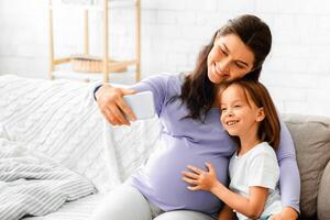 Happy expecting woman taking selfie with her little daughter while sitting on sofa at home, free space photo