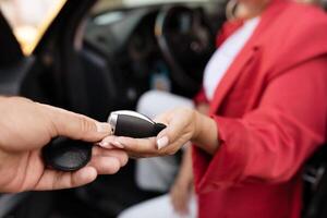 A man extends a set of car keys to a woman wearing a red jacket, who is seated inside a vehicle. The interaction suggests a transfer of ownership or a lending arrangement. photo