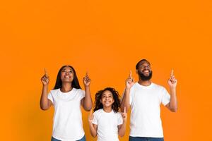 Portrait Of Excited Black Family Of Three Looking And Pointing Two Fingers Up Above Their Heads At Copy Space Isolated On Yellow Orange Background, Demonstrating Free Place For Your Advert, Banner photo