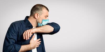 Sick Man Coughing Into Elbow Wearing Protective Medical Mask Having Coronavirus Pneumonia Standing Over Gray Background. Panorama photo