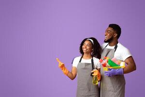 Cleaning Service Ad. Portrait of smiling black couple holding basket with detergents and pointing aside at copy space on pink background, panorama photo