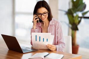 Business, Technology And Communication Concept. Young woman making phone call while looking at document with graphs photo