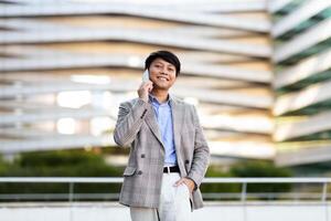 A young Asian man stands confidently in a tailored suit while engaging in a phone conversation outside a contemporary architectural structure during daylight. photo