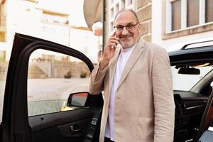 A well-dressed man stands beside his parked car in an urban setting, smiling while engaged in a phone conversation. The environment reflects a charming city atmosphere. photo