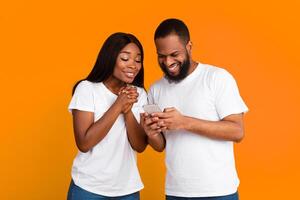 Excited black couple looking at cell phone screen, using new cool application, standing isolated on orange studio wall. Joyful African American man and woman using smartphone together, sharing media photo