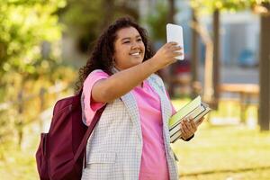 A young student with curly hair smiles brightly while taking a selfie outside. She holds a stack of books and carries a backpack, enjoying a sunny day in a vibrant environment. photo