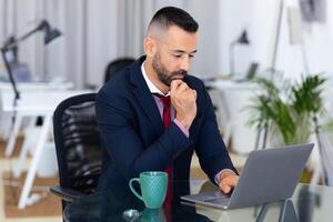 A man in a suit is focused on his laptop in a contemporary office. He appears to be deep in thought, holding his chin while typing, surrounded by various office elements. photo