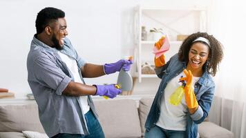 Cheerful african american couple playing with sprayers while cleaning flat together, having fun at home, free space photo