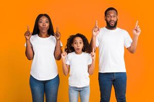 Wow, Cool Offer. Portrait Of Surpised African American Family Of Three People Standing On Yellow Orange Background Pointing Fingers Up Showing Free Copy Space And Empty Place For Advertising photo