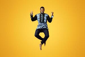 Emotional black guy showing happiness over pink studio background. Smiling african american man in colorful traditional clothes jumping up and posing, raising fists up, expressing emotions photo