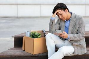 A jobless Asian man in a gray blazer and white pants sits on a wooden bench. He has his belongings in a cardboard box beside him. He looks down at his phone with a worried expression. photo