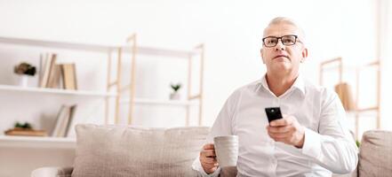 Smart Tv. Concentrated Senior Man Holding Remote Control And Coffee Mug, Empty Space, Low Angle View photo