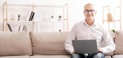 Smiling Mature Man Holding Computer On His Lap, Sitting On Couch, Typing On Keyboard And Looking At Camera, Copyspace photo