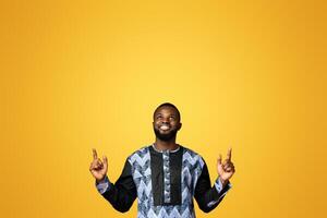 Happy black guy in african costume pointing up at free space, showing advertisement on pink studio background. Positive african american man looking up, directing at empty space for text photo