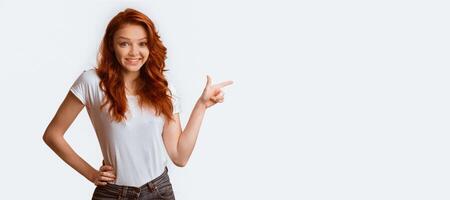 Cheerful Red-Haired Girl Pointing Finger Aside Posing Over White Studio Background. Panorama, Empty Space For Text photo