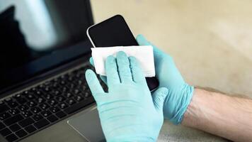 Unrecognizable Man Disinfecting Smartphone Before Use Sitting At Workplace, Wearing Protective Gloves Indoor. Coronavirus Protection And Hygiene. Cropped, Selective Focus photo
