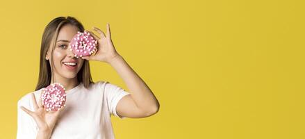 Portrait of funny positive woman holding donuts near her face and sincerely smiling while posing over yellow background in studio, empty space photo