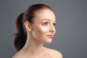 Pretty Young Lady With Heart-Shaped Moisturizer Cream On Cheek Caring For Face Posing Looking Aside On Gray Studio Background. Skin Care Routine, Cosmetics Advertisement. Panorama With Empty Space photo