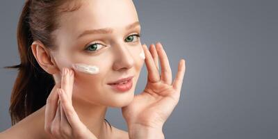Closeup Of Red-Haired Young Woman Applying Moisturizing Cream On Face Caring For Skin Smiling To Camera Standing Posing On White Background. Studio Shot. Skincare For Young Lady photo
