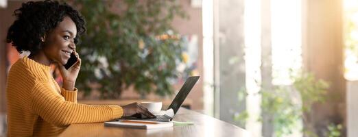 Smiling young black woman talking to clients by phone, working online in cafe with laptop, copy space, side view photo