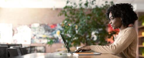 Young african woman working in cafe, using laptop, side view, empty space photo
