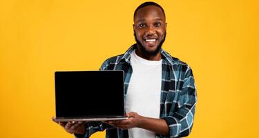 Smiling African Guy Showing Laptop Computer Blank Screen To Camera Recommending Great Website Standing On Yellow Studio Background. Online Service Advertisement. Mockup photo