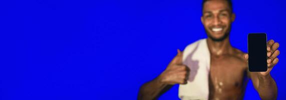 Workout Application. Athlete Guy Showing Smartphone Blank Screen Gesturing Thumbs-Up Standing On Blue Studio Background. Mockup, Shallow Depth photo