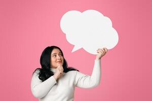 Pensive plus size woman holding and looking at blank speech bubble over her head, standing on pink background photo