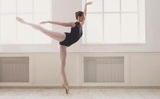 Classical Ballet dancer side view. Beautiful graceful ballerine in black practice arabesque ballet position near large window in light hall. Ballet class training, high-key soft toning. photo