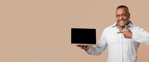 Awesome website. Middle aged positive afro man pointing at blank screen of laptop, orange studio background photo