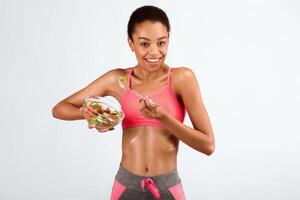 Diet And Fitness. Slim Afro Woman Eating Vegetable Salad Smiling At Camera Standing Over Pink Studio Background. photo