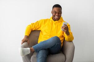 Cheerful Black Millennial Man Using Cellphone Browsing Internet And Texting Sitting In Armchair Over Gray Wall Indoors. People And Gadgets. Mobile Communication Concept photo