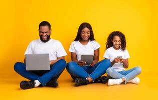 Family Gadgets. Portrait of happy African American parents and their little daughter holding and using different electronic devices while sitting on the floor over yellow studio background photo