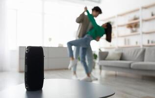 Millennial Asian couple dancing to popular music at home, selective focus on portable wireless speaker on table, copy space. Young family moving to their favorite song, using modern device photo